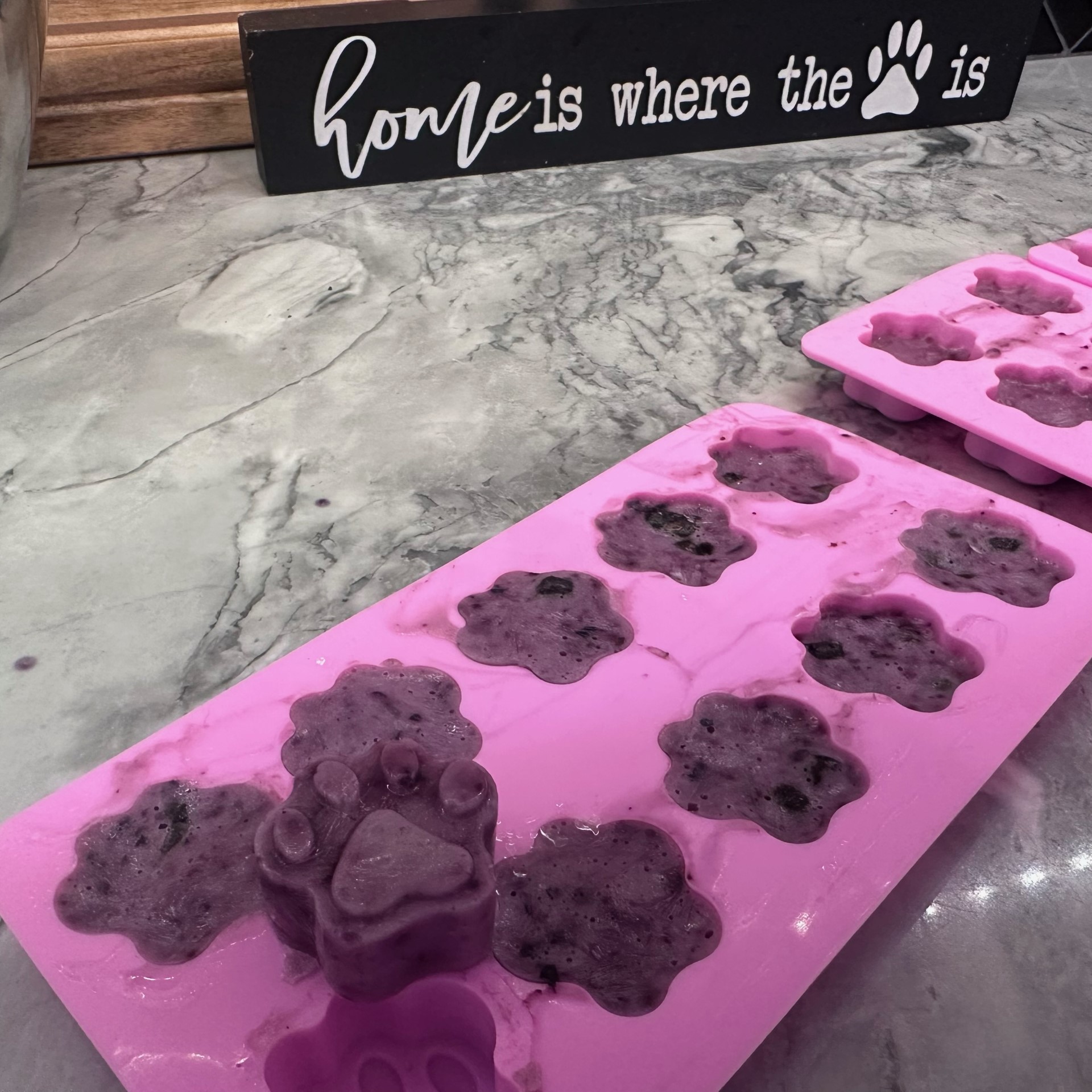 Pink silicone mold filled with frozen dog treats in the shape of paw prints, placed on a marble countertop with a decorative sign that reads 'home is where the paw is.'